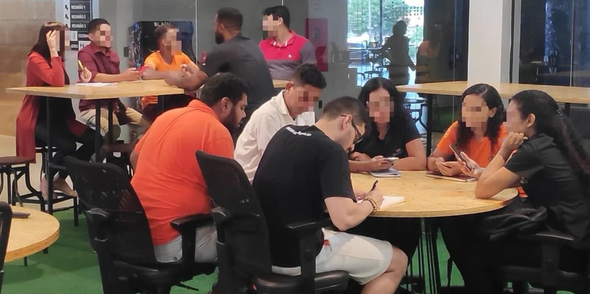 People seated around tables in an office lounge, discussing and taking notes.